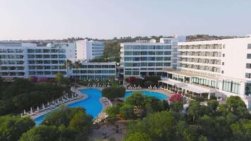 Aerial view of a resort in Ayia Napa, Cyprus, with a large pool, modern white buildings, lush greenery, and gradual panning motion in soft sunlight. video