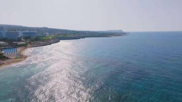 Sweeping aerial view of Ayia Napa, Cyprus, showcasing rugged cliffs, sparkling Mediterranean waters, and a resort town with scattered greenery. video