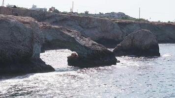 Rugged cliffs and clear blue waters in Ayia Napa, Cyprus. Gentle waves lap against the shore with palm trees and white buildings in the background. video