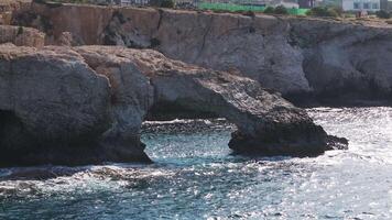 Aerial pan of Ayia Napa's limestone cliffs, natural arches, and caves with turquoise waters and a resort in the background. Waves gently move below. video
