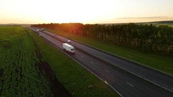 Aerial shot of the highway in the rays of the setting sun and a moving truck with a semi-trailer. Heavy truck driving on an asphalt road along green fields. Top view video