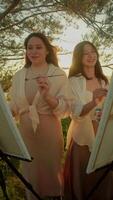 Two young students are engaged in conversation about their paintings, which they are creating in midst of picturesque summer setting, with warm sunset in background. Portrait. video