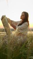 Charming artist enjoys creating works of art on canvas. She paints with watercolor paints in of summer meadow surrounded by tall grass, against background of setting sun. Portrait. video