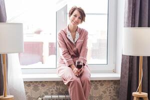 Beautiful woman wears pajamas with a glass of red wine sits on the windowsill near beautiful window at home. photo
