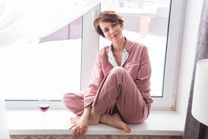 Beautiful woman wears pajamas with a glass of red wine sits on windowsill near beautiful window at home photo