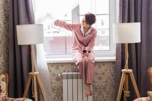 Beautiful woman wears pajamas with a glass of red wine sits on the windowsill near the beautiful window at home. photo