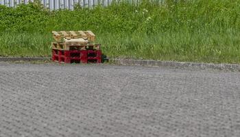 Red and pure wooden pallets are stacked on a driveway in front of a green area, the foreground and paved photo