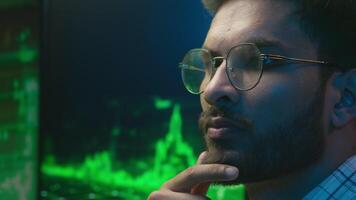 Indian Arabian man in glasses thinking contemplate ponder solving coder trader IT specialist male ethnic guy businessman business programmer analysing trading chart graph crypto currency neon indoor video