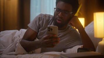 Disappointed African American ethnic man guy client male client use mobile phone smartphone reading bad news frustrated user lying bed sadness fail losing problems no connection at home bedroom night video
