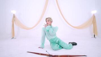 A person in a mint green outfit poses elegantly on a white backdrop with draped fabric. The setting is bright and minimalistic video