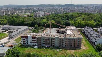 Aerial view of residential building under construction. Multi-story apartment complex being built near a forest edge, with cranes and urban skyline in the video