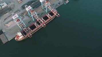 Aerial view of a cargo ship docked at an industrial port with cranes unloading bulk materials into ship holds video