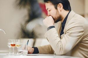 Close up Photo of Thoughtful Sommelier Making Notes in Degustation Card during passes Exam on Alcoholic Beverages Experts Occupational sitting Front of Glass with Various Spirits