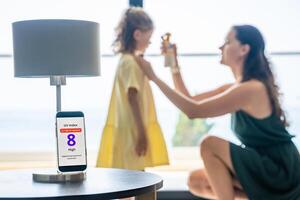 Smartphone with high UV alert in focus, while mother applies sunscreen to daughter in background. Real-life response to sun safety warning. High quality photo