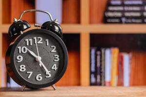 A Black alarm clock on wooden table with bookshelf in the background photo