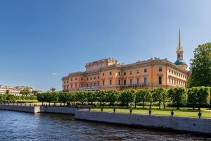 Mikhailovsky Castle with a golden spire stands by the river, with even trees growing along the bank. photo
