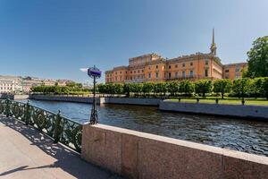 Mikhailovsky Castle on the river bank, with a bridge featuring a decorative fence, under a clear blue sky. photo