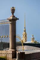 A view of the Peter and Paul Cathedral spire and a bridge over the river, framed by a forged grille and column. photo