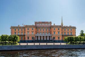 Mikhailovsky Castle of pink color with columns and a spire, stands on the river bank under a clear sky. photo