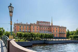 Mikhailovsky Castle, illuminated by the sun, on the river bank, with a bridge and a lantern in the foreground. photo