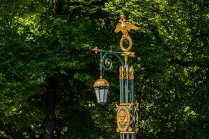 A decorative street lamp with a golden double-headed eagle and a lantern, against a background of green tree foliage. photo