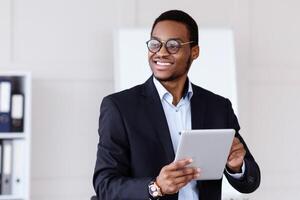 Modern gadgets and business. Joyful african american young businessman in suit using digital tablet in office, working online, getting ready for presentation, looking at copy space and smiling photo