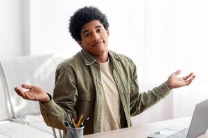 Puzzled black student sitting in front of laptop, not understanding difficult lesson, having difficulty studying online. African American teen guy shrugging shoulders, feeling confused during exam photo