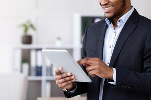 Smiling black manager in suit using digital tablet, office interior, copy space. Happy african american businessman using business application on pad, enjoying user friendly software photo