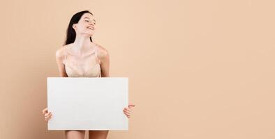Smiling young female with vitiligo skin disorder holding white blank placard, beautiful millennial lady standing in underwear over beige background in studio, enjoying her different beauty, copy space photo