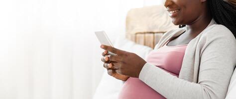 Joyful Black Pregnant Lady Spending Time With Smartphone At Home, Browsing Internet Or Messaging With Friends, African Expectant Woman Relaxing On Comfortable Bed, Side View Shot With Copy Space photo