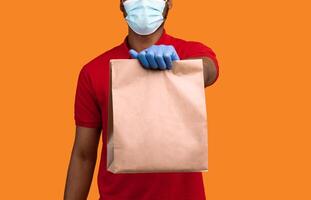 Delivery During Pandemic. Close up of african courier wearing surgical mask, protective gloves and red uniform holding and giving craft paper package with blank space for design, orange background photo