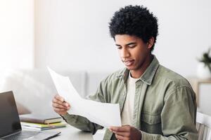 Black teenager studying remotely on laptop, working on coursework, coordinating his school project with tutor at home. African American student with papers using PC with empty screen, mockup photo