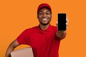 Delivery Service Application. Smiling african american man showing smartphone with blank screen for mockup, wearing red cap, uniform and thermo backpack bag, holding box, isolated on orange background photo