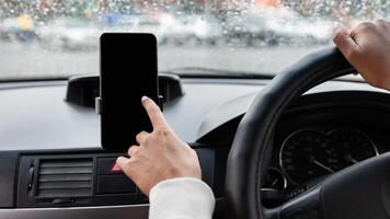 Online map and loud call at driving in city. Millennial african american woman in car, holds steering wheel and uses phone with blank screen, looks for route, view of windshield with raindrops, pov photo