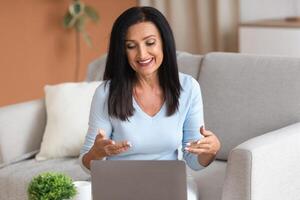 Online Call And Communication. Portrait of smiling mature woman sitting on couch at table, using laptop and having virtual conference on computer, talking to webcam, explaining plan photo