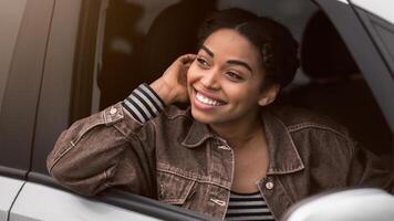 Pleasant looks african american female with glad expression, being satisfied with unforgettable journey by car, sits on driver s seat, enjoys music and look at open window. People, driving, transport photo