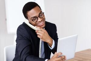 Happy black man in suit office manager or businessman talking on phone, using digital tablet at workdesk, consulting clients, using modern technologies, copy space, office interior photo