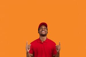 Portrait of optimistic african american delivery man in red t-shirt and cap smiling, looking and and pointing fingers up on copy space text or product isolated over orange studio background photo