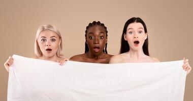 Three Emotional Multi-Ethnic Women Hiding Behind Bath Towel With Shocked Face Expression, Group Of Dazed Ladies Standing With Open Mouth Over Beige Background, Reacting To Amazing Offer, Copy Space photo