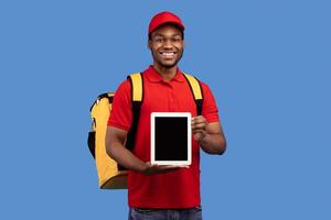 Modern Technology And Delivery. Smiling black male courier holding digital tablet with empty screen for mockup and showing to camera, wearing red uniform and yellow thermo bag, standing at blue studio photo