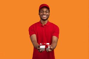 This Present Is For You. Front view portrait of smiling black delivery man holding wrapped gift box giving it to the camera, isolated on orange studio background, greeting with special day photo