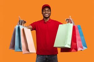 Fast Shipment Concept. Cheerful black deliveryman wearing red uniform and cap holding and showing colorful shopping bags with orders and presents, isolated over yellow orange studio background photo