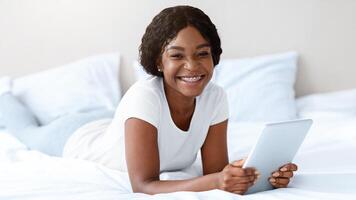 Pretty black woman with digital tablet smiling at camera, enjoying new useful applications for pad, reading book or watching something online, surfing on social media, bedroom interior, copy space photo
