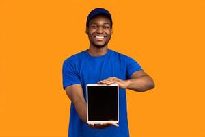 Delivery Application. African american courier man holding digital tablet with blank screen for mock up and showing it to camera, wearing blue cap and uniform, standing isolated on orange background photo