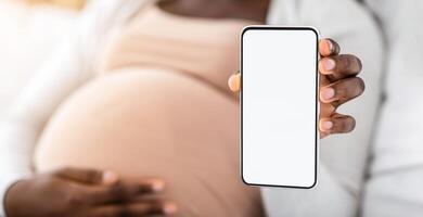 Pregnancy App. Black pregnant lady demonstrating smartphone with blank white screen, recommending new app for expecting women, demonstrating maternity shopping website, mockup image with copy space photo