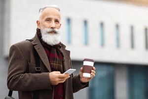Handsome aged grey-haierd man holding paper cup with coffee and using modern smartphone, walking by street during break, copy space. Elderly man going to job, drinking morning coffee photo