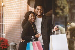 Fun walk for shopping in November and final sale. Cheerful young african american guy and lady in coats hug and look at camera, hold colored bags with purchases, near brand store, outdoor, copy space photo