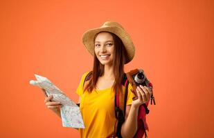 Happy young woman backpacker with camera holding city map over yellow studio background. Joyful lady traveller in summer hat looking at camera and smiling, going for sightseeing abroad, copy space photo