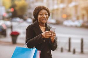 Message about start of sales season and Black Friday for shopaholic at mobile app. Happy millennial african american woman holding colored bags, smartphone and ready for shopping, outdoor, free space photo