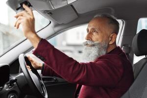 Aged grey-haired man driver fixing mirror in car before moving. Elderly businessman in stylish suit getting ready before travelling by his auto, setting rearview mirror, empty space photo